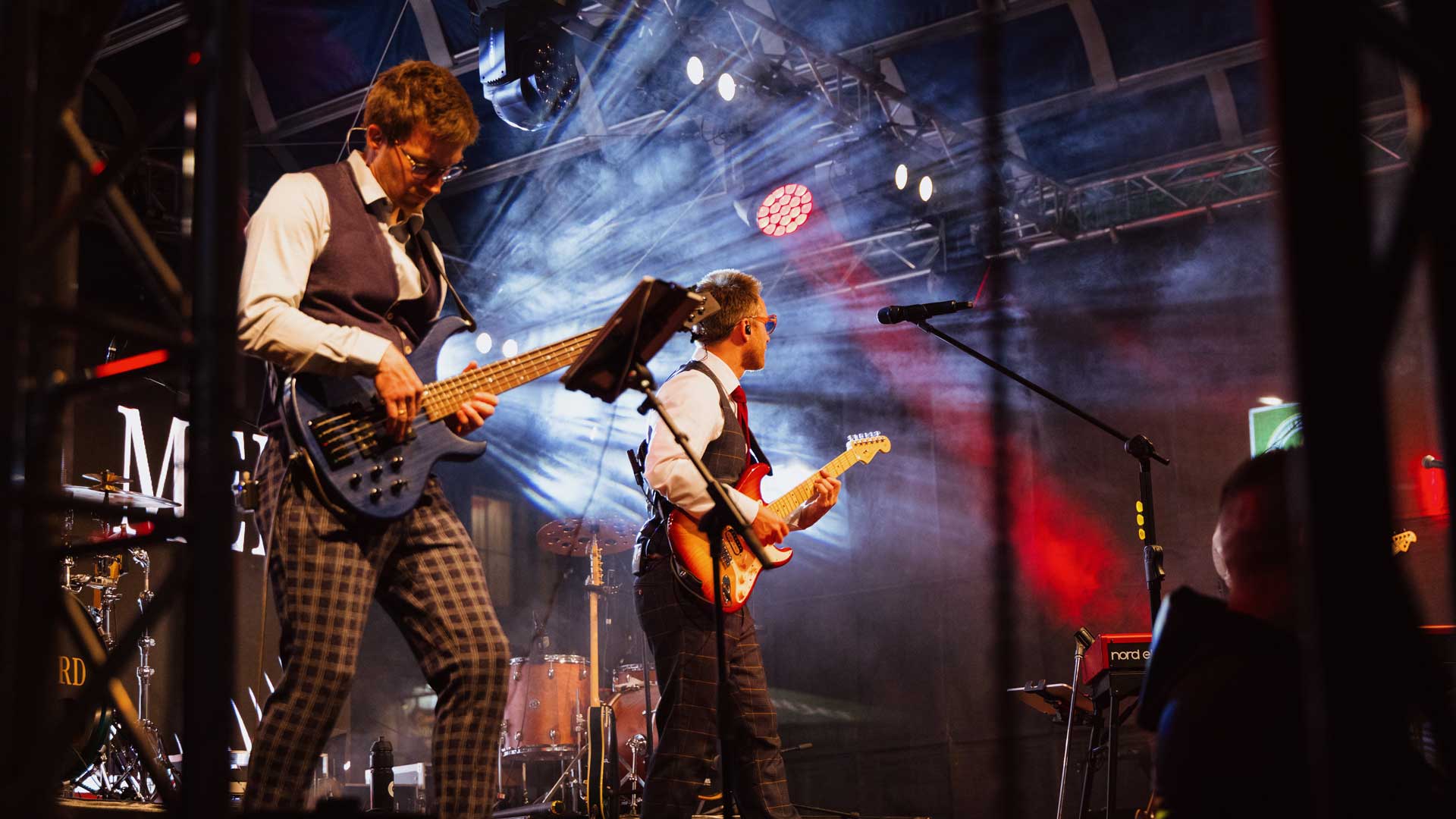 Zwei Musiker in Westen und mit Brillen spielen auf der Bühne unter bunten Lichtern E-Gitarre, während im Hintergrund bei einem Live-Konzert Schlagzeug und Mikrofone zu sehen sind.